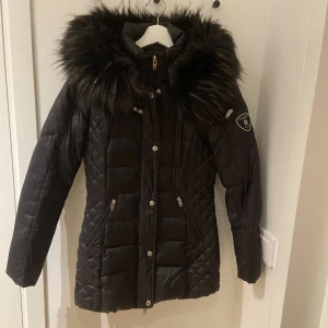 Rockandblue beam mid jacket - Beam Mid är en kort dunjacka i en elegant, figurnära modell. Jackan har en tvåvägsdragkedja och en huva med fuskpälskant som går att knäppa av och på. Jackan är behandlad med vattenavvisande BIONIC-FINISH®ECO.