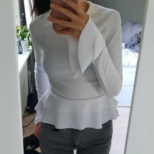 Vit blus - Vit långärmad blus med volanger. Jag säljer den pga att den inte kommer till användning och är endast testad. Det finns några små fläckar på ena armen som var när jag gick hem den, men dem syns knappt när man har den på sig. Köparen står för frakten!