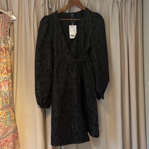 Svart klänning med knytning, puffärm & mönster  - En svart klänning med v-ringning, knytningar vid sidan & puffärm. Helt ny med prislappen kvar. Var tanken att jag skulle ha den på nyår men blev aldrig använd. Storlek: Xs!  Nypris: 500.  Kontakta mig vid frågor eller intresse.