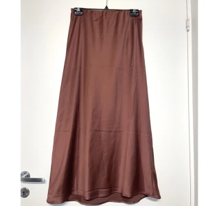 Vadlång Kjol - Vadlång kjol i satin imitation (polyester). Färgen är mörk magenta/röd-lila, (färgen gör sig ej tillrätta på bilderna). Jag är 172cm för referens på längden. Skick: aldrig använd, dock tvättad några gånger. Nypris ca 150kr.
