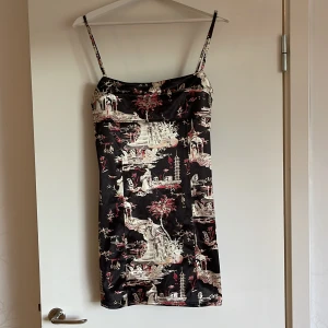 Kort klänning - En kort svart klänning med vitt och rött mönster. Åtsittande passform. Glansigt tyg. Justerbara axelband. Storlek M.