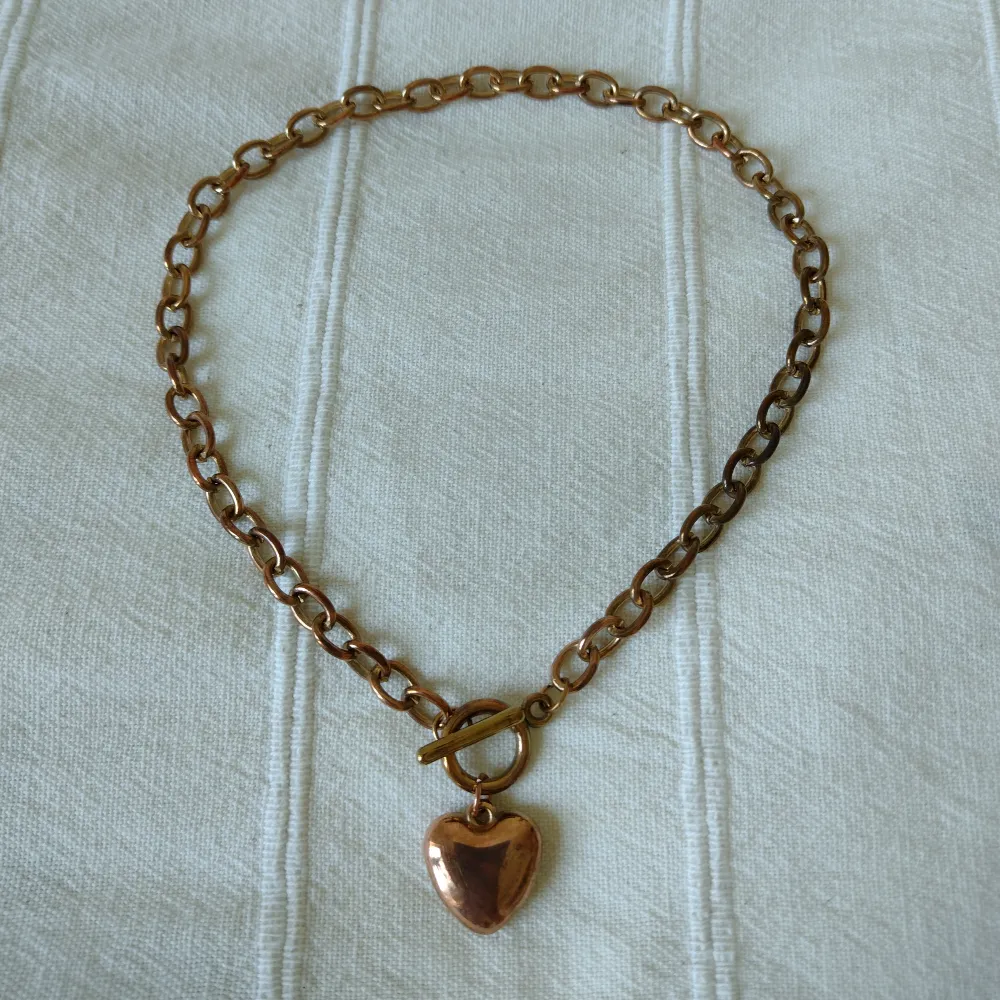 Halsband i metall med berlock i form av ett hjärta. Ca 40 cm långt. Mässings-/kopparfärgat - färgen skiftar lite. Kan skickas med ett frimärke (18 kr).. Accessoarer.