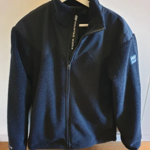 Unisex Hybrid Pile Fleece! - Unisex Hybrid Pile Fleece från Mountain Works. Varm och mjuk pilefleece i unisexmodell, tillverkad i 100 % återvunna material.  Fleecen har ett värmande yttertyg och ett vattentätt tyg på bröst och axlar som skyddar mot regn och vind. Aldrig använd.
