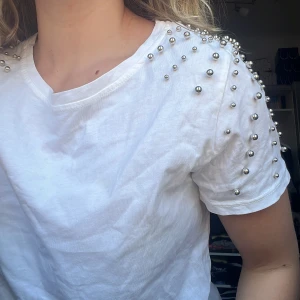 Vit T-shirt med pärlor  - En Vit T-shirt med stora silvriga pärlor på axlarna som går både framåt och bakåt på axlarna, aldrig använd. Står ej storlek men skulle gissa att den är i M
