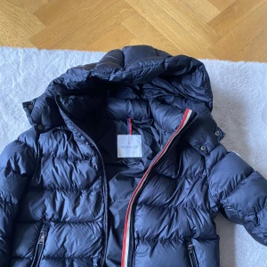 Moncler dunjacka stlk 152  - Tjock dunjacka från Moncler. Stlk 152 (12år) men lite mindre i storlek.  Se bild på små hål på ena ärmen. En flaws på ryggen  Inköpt på NK, bild på QR kod finns.  Nypris idag ca 7000kr 