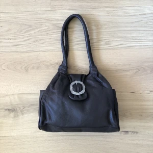 Brun läderväska spänne - Superfin brun läderväska med ett silvrigt spänne. B34,H26,D10 handtag ca 50 cm. Blå/vitt rutigt innertyg. 