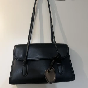 Svart axelväska - En helt ny svart axelväska. Stängs med två magnetknappar under ”flärpen”, finns även ett litet fack inuti väskan som försluts med dragkedja. 