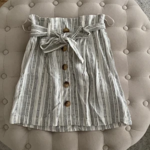 Kort kjol - Kjol från Topshop, storlek 32, aldrig använd med prislapp kvar. Vit med grå linjera, tyget är som linnetyg, knappar på framsidan som kan öppnas hela vägen.