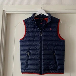 Väst Polo Ralph Lauren - Blå/röd dunväst i använt men bra skick. Storlek junior L (14-16) men tycker den är ganska liten i storleken så passar 150-164 cm. Påse till västen medföljer.