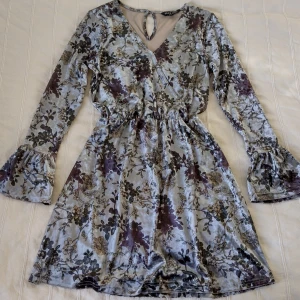 Silvergråblå klänning från Brave Soul - sammet/velour - storlek S - Mönstrad klänning i något sammetsliknande material. Omlottmodell upptill och resår i midjan. Fina volanger vid ärmsluten. Fin silvergrå/blåaktig färg med blommönster. 95% polyester 5% elastan. Storlek S. Bara använd en gång.