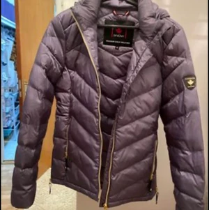 Dunjacka Snow str38 - Stålblå lättviktsdunjacka från Canada Snow i trendigt glansigt material. Jackan är fodrad med 50% dun och 50% polyestervadd. Jackan är lätt figursydd och har lyxiga i guldfärgade detaljer, avtagbar huva, dolda muddar i ärmslut.Använd endast ett fåtal gång