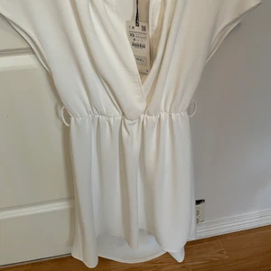 Vit oanvänd klänning  - Oanvänd vit klänning köpt på zara. Kommer med tillhörande brunt läder (fejk) bälte med guld detalj. Perfekt klänning till examen eller student. Klänningen är lite längre nertill och har en fin urringning. Passar även en storlek S 