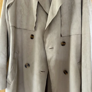 Trench coat - En beige lite tunnare trench coat i mocka. Köpt i polen. Helt oanvänd. Skärp medföljer. Står att det är XL men passar M-L