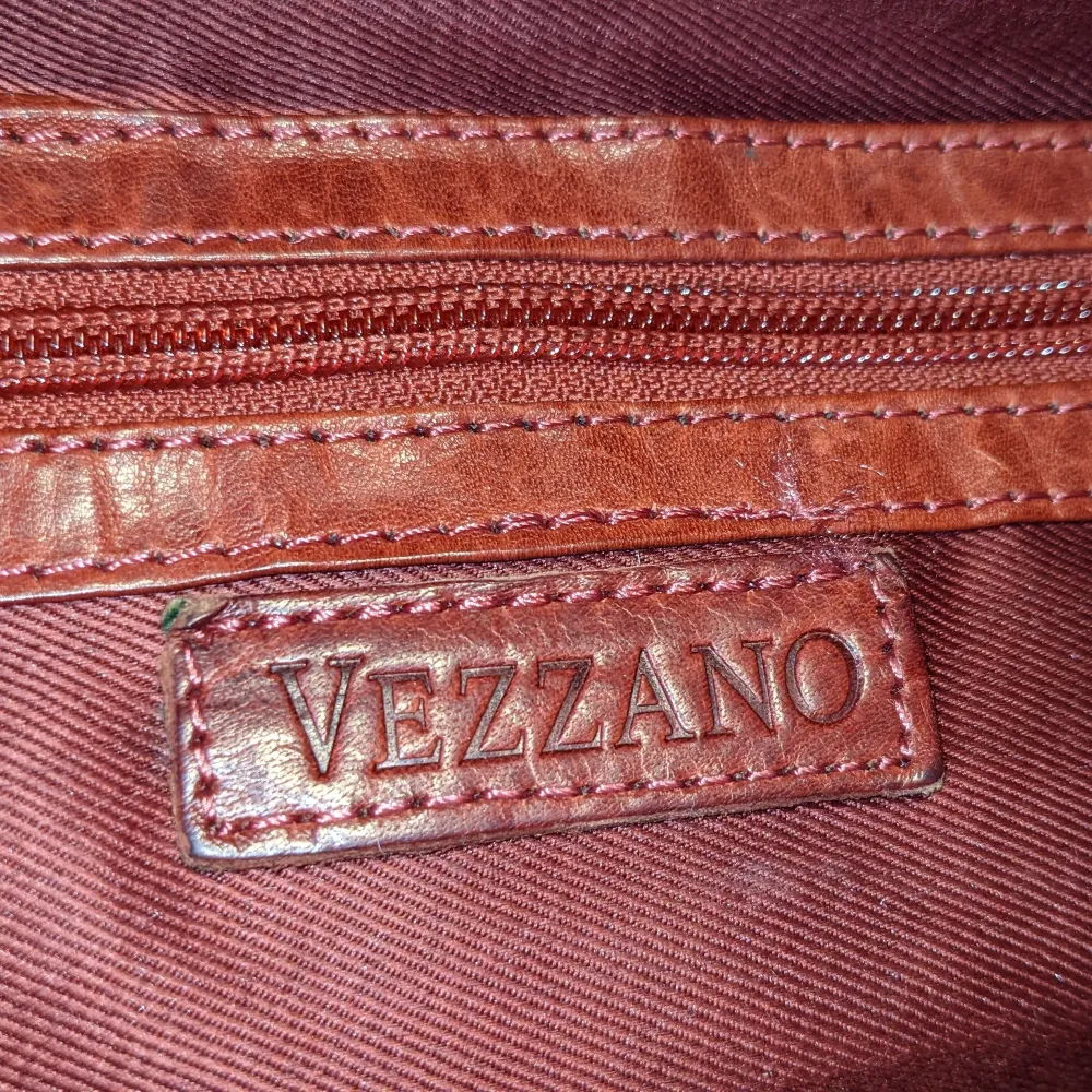 Rymlig och trevlig väska i skinn. Märket är Vezzano och den har en brunröd färg. Går att bära med både den långa remmen och som en handväska med de korta. Rymmer mycket och har små fack på insidan. I gott skick!. Väskor.