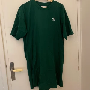 Adidas T-shirt klänning - En t-shirtklänning från adidas. Ett broderat adidasmärke på bröstet på framsidan och på ryggen ett stort tryck. Jag är 170 och den går till mitten på låret på mig. Storlek S men är oversized