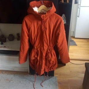 Höst Jacka (Indiska) - Orange-brandröd lång fodrad Jacka.  4st reglerbara snören för åtsittande eller avslappnad look, 2 fickor. Använd 1 gång , köpt 2019  Nypriset var 800:-  Ps. Medhörande päls till luvan hittas ej. Jackan framhävs mer orange på bild än i verkligheten.