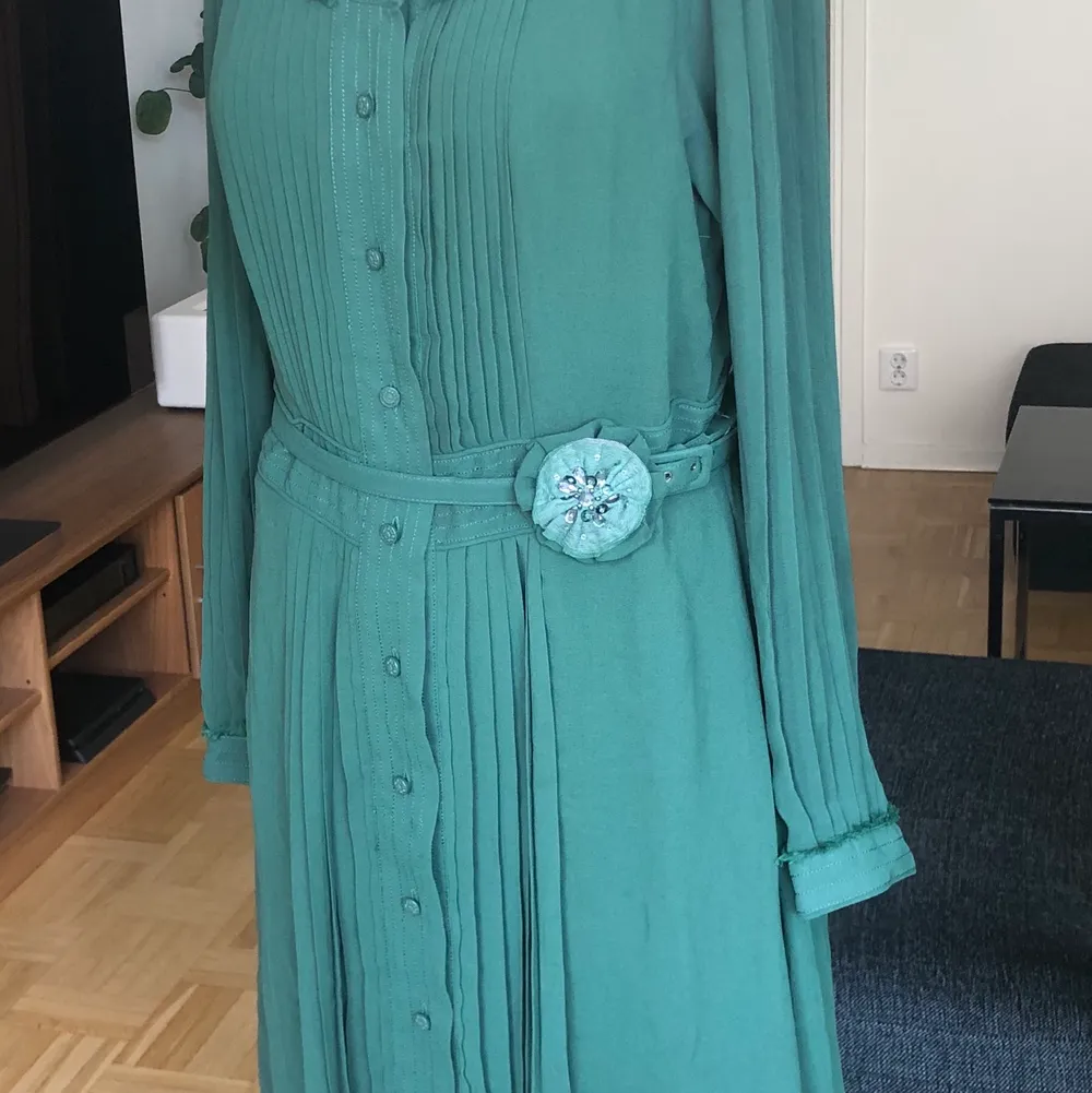 Grön plisserad klänning med skärp i midjan och en tillhörande brosch i form av en blomma. Sparsamt använd.. Klänningar.