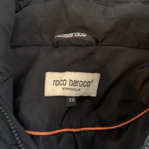 Dunjacka Roco baroco  - Det är en svart dunjacka med ej äkta pälskrage. Ganska lång, går precis nedanför rumpan. Knappt använd och i mycket fint skick. Köparen betalar för frakt. 