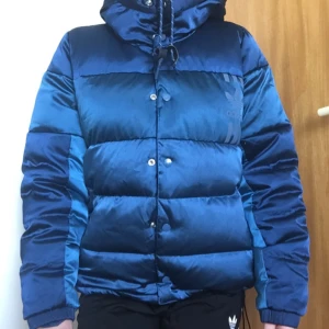  - Finaste dunjackan från Adidas! I två blåa, silkiga, nyanser. Dragsko i botten och mudd på insidan. Fickor med dragkedja. Luva och dragsko i halsen. Knäpps med knappar och dragkedja. Världens finaste jacka!  