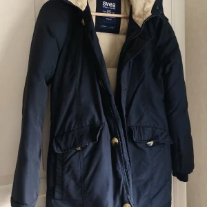 Vinterjacka från SVEA - Marinblå vinterjacka med beiget innerfoder i en lite längre modell från SVEA (bomull, nylon, dun, fjäder). Storlek XS, men passar även S. Sparsamt använd.