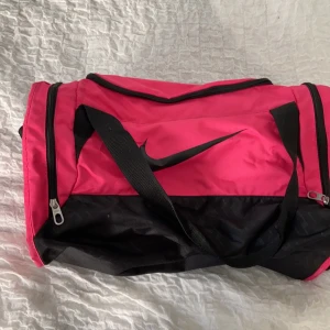 Väska Nike  - En träningsväskan från Nike med två fack på sidorna för skor tex och även ett stort i mitten. Två kortare band och ett långt axelband. Rosa och svart. Möts upp i Stockholm, kan också frakta men isåfall står du själv för frakten. 