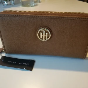  - Modell: Honey Za Wallet  Ny, oanvänd med etikett på. Köpt i USA. Färg : Brun med detaljer i guldfärg.  Gratis leverans i söderort.  Mått: Höjd : 10 cm Längd/Bredd : 20 cm  Produktinformation: Dragkedja i full längd Fack för kreditkort Myntficka med dragkedja Dragspelsfack       