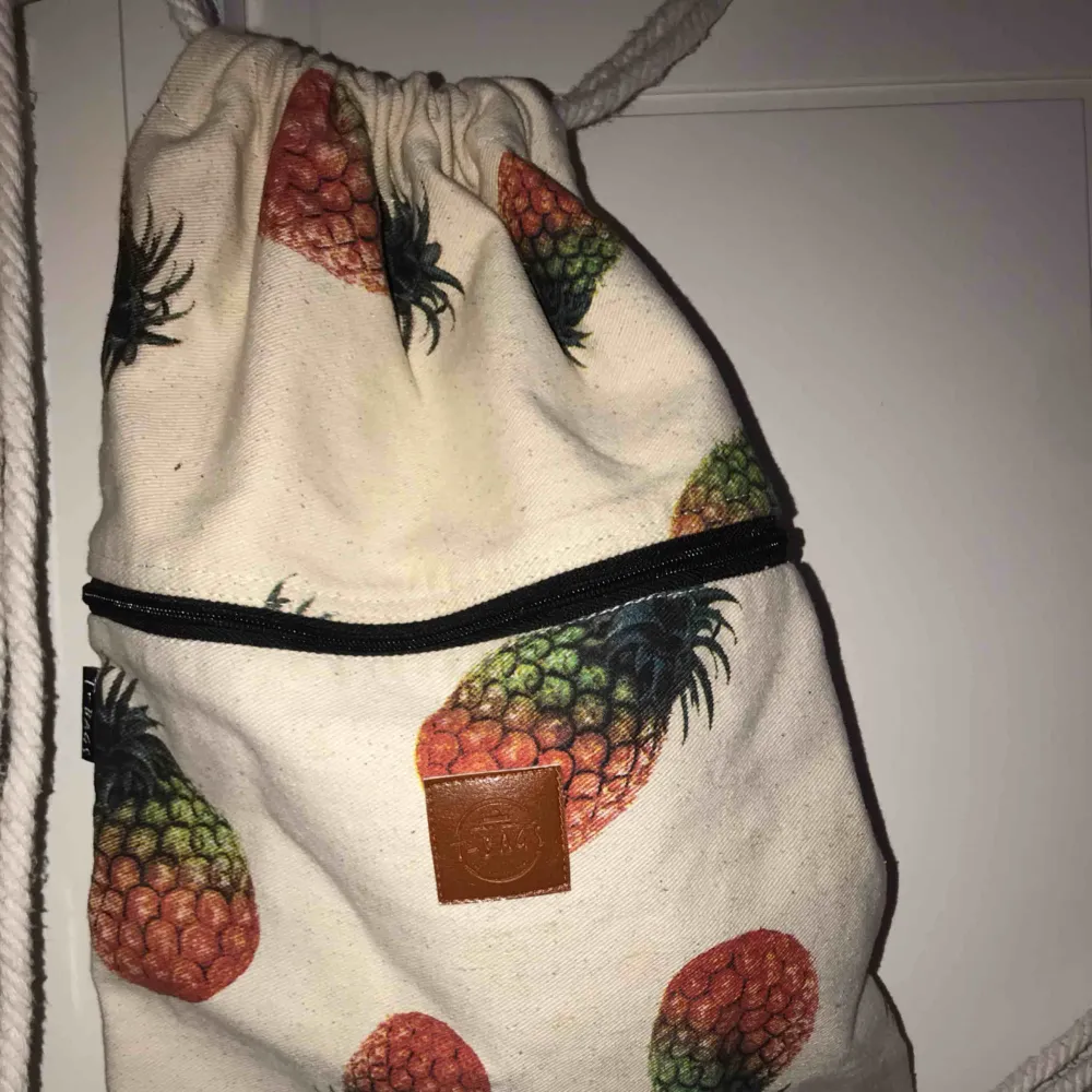 En vit väska med tryck av ananaser, den har snören/rep som man knyter ihop och bär den med. Väskor.