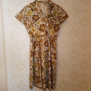  - Klänning från 60-talet med gulbrunt mönster på vit bakgrund. En liten jacka i samma tyg, som kan stängas med två små dolda tryckknappar, ingår. Jättefint skick på bägge! Frakt ingår.