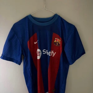 FC Barcelona Lewandowski matchtröja - Säljer en officiell FC Barcelona hemmatröja med Lewandowski 9 på ryggen. Tröjan är blå med röda ränder, har klubbmärke och Nike-logga på bröstet samt sponsortryck. Kortärmad modell i lätt och ventilerande material, perfekt för fotboll eller supporterliv.