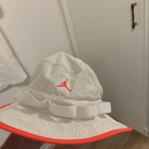Jordan vit bucket hat med neonröd kant - Bucket hat från Jordan i vit bomull med neonröd kant och broderad Jumpman-logga framtill. Hatten har bred brätte, justerbart band och coolt svart innerfoder med textdetaljer. Perfekt för att sticka ut i sommarvärmen.
