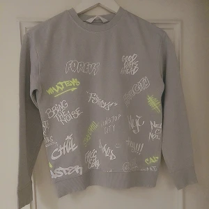 Grå sweatshirt med graffiti-tryck, storlek 10 år - Säljer en grå sweatshirt med coola graffiti-inspirerade tryck i vitt och neongult över hela framsidan. Tröjan har rund hals, långa ärmar och relaxed fit. Perfekt för dig som gillar streetstyle och vill sticka ut lite extra.