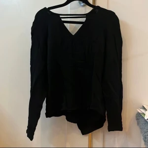 Svart blus från Tiger of Sweden - Säljer en svart blus från Tiger of Sweden med v-ringning och snygga banddetaljer vid halsen. Blusen har långa ärmar och en stilren, minimalistisk look som passar perfekt till jeans eller kjol.