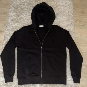 Svart hoodie från These Glory Days - Svart hoodie med dragkedja från These Glory Days. Klassisk modell med huva, snörning och känguruficka framtill. Tillverkad i mjuk bomull och har långa ärmar. Perfekt för chill dagar eller när du vill ha en enkel och stilren look.