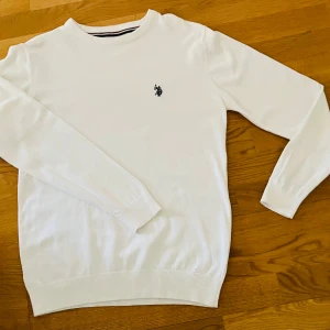 Vit stickad tröja från Polo - Vit tröja från Polo. med rund halsringning och broderad logga på bröstet. Tröjan har långa ärmar och ribbade muddar vid ärmslut och nederkant. Perfekt för en clean och stilren look.