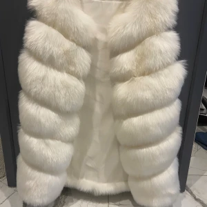 Vit fluffig pälsväst - Supermjuk och lyxig vit pälsväst med breda, horisontella paneler. Västen är lång och har en riktigt fluffig look som ger en exklusiv känsla. Perfekt för dig som vill sticka ut och hålla värmen med stil.