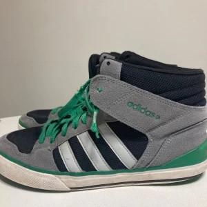 Adidas sneakers med gröna detaljer - Snygga Adidas sneakers med högt skaft i svart mesh och grå mocka. Klassiska vita ränder på sidan och gröna snören samt gröna detaljer på sulan. Perfekt för dig som gillar streetstyle och vill sticka ut med färg. Skorna har rund tå och platt sula.