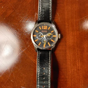Svart & orange klocka från Hugo Boss - Snygg klocka från Hugo Boss Orange med svart urtavla och orange detaljer. Klockan har ett robust rostfritt stålboett, svart läderarmband med vit söm och tydliga orange index. Urtavlan har mönstrad bakgrund och flera små visare för dag och datum. Perfekt för dig som gillar sportig stil.