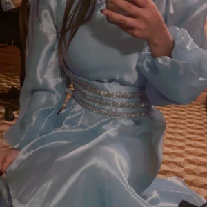 Ljusblå långklänning med glitterdetaljer - Säljer en ljusblå långklänning med glansigt tyg och puffiga långärmar. Midjan är dekorerad med tre glittriga silverband som ger en extra lyxig känsla. Klänningen har en elegant och flytande siluett, perfekt för dig som vill sticka ut med stil.