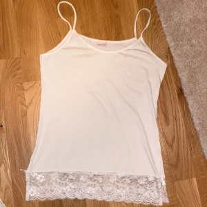 Vitt linne med spets - Vitt linne från Fil Rouge med tunna axelband och bred spetskant nertill. Spetsen har blommigt mönster och en liten rosett på båda sidorna för extra detalj. Liten sminkfläck på framsidan som knappt syns och säkert går att tvätta bort. Storleken står inte men skulle säga S/M