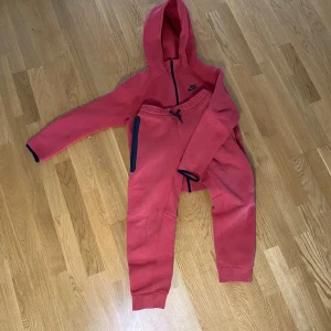 Röd Nike hoodie i fleece och byxor fleecen är XL (158-170 cm) och byxorna är L (147-158 cm) - Nike hoodie i en snygg röd färg med svart logga och dragkedja. Nike byxor i snygg röd färg med svart logga på benet.Tröjan har huva, långa ärmar och är gjord i mjuk fleece. Perfekt för chill eller träning. Matchande byxor medföljer, båda med svarta detaljer och dragsko i midjan.
