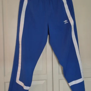 Blå Adidas träningsbyxor med vita ränder - Snygga blå träningsbyxor från Adidas i retro stil med klassiska vita ränder längs sidorna och runt bensluten. Byxorna har elastisk midja och Adidas logga på framsidan. I nyskick.