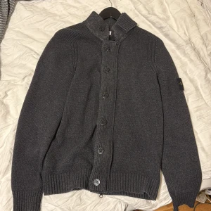 Mörkgrå kofta från Stone Island - Säljer en mörkgrå stickad kofta från Stone Island med ribbade muddar, hög krage och både dragkedja och knappar framtill. Klassisk logga på ärmen. Perfekt för lager-på-lager under höst och vinter.