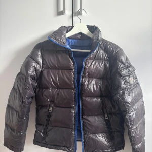 Moncler jacka - Så vacker och cool moncler jacka. Den är svart på utsidan och blå på insidan, dragkedjan är även blå. Den är köpt för länge sen så den har tyvärr inte en qr kod.   Några få defekter är att färgen på knapparna har slitits ut och logan på dragkedjan (se bild 7). En till defekt är att cartoonen sitter löst eftersom sömmarna är lösa. På bild 9 ser man 5 pytte små ”hål” men inget som man tänker på.  Storleken är 152cm men den passar mig som bär XS jätte fint! Modellen på jackan är att den ska vara li