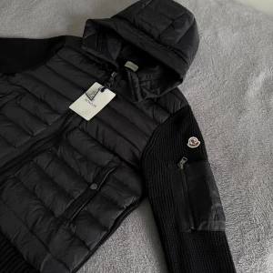 Moncler cardigan  - Svart cardigan från Moncler med huva, stickade ärmar och detaljerad med dragkedja på ärmen. Jackan har quiltad front och ribbade muddar. Perfekt för dig som vill ha en stilren och sportig look med premiumkänsla.