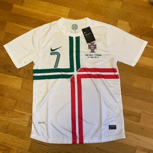 Portugal Ronaldo 7 Nike fotbollströja - Säljer en vit Portugal fotbollströja från Nike med gröna och röda korsande linjer på framsidan, nummer 7 och Ronaldo på ryggen. Tröjan har korta ärmar och är tillverkad i lätt Dri-FIT material som andas. Landslagsmärket och matchdatumet 27/06/2012 finns tryckt på bröstet.