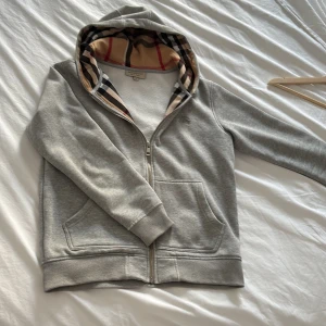 Grå hoodie från Burberry med dragkedja - Grå hoodie från Burberry med klassiskt rutigt foder i huvan och broderad logga på bröstet. Tröjan har dragkedja framtill, kängurufickor och ribbade muddar. Perfekt för dig som gillar stilren design med lyxig touch.