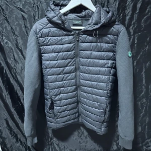 Svart cardigan jacka Save The Duck  - Svart cardigan jacka från Save The Duck med quiltad framsida och huva i syntetmaterial. Ärmar och rygg är stickade för en skön kontrast. Jackan har dragkedja framtill och två fickor med dragkedja. Perfekt för dig som gillar stilren och funktionell design. Jackan har som man ser på bild 3 och 5 små defekter, lite av vitt tryck på insidan faller av och lite nopprig, men den är knappt använd och är i superbra kvalitet. Den är i storlek S hör av dig vid frågor och pris kan diskuteras vid snabb affär