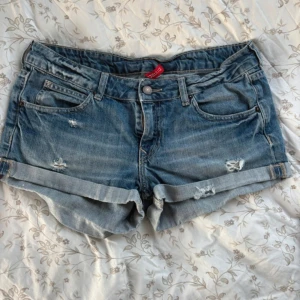 Blå Lågmidjade jeansshorts med slitningar - Säljer ett par klassiska blå jeansshorts från Gina Tricot med snygga slitningar och upprullade benslut. Shortsen har fem fickor, knapp och dragkedja framtill samt en skön midwaist-passform. Perfekta för sommardagar!