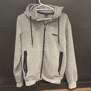 Grå hoodie med dragkedja från BOSS - Snygg grå hoodie från BOSS med svart broderad logga på bröstet. Tröjan har hel dragkedja, två fickor med svarta dragkedjor och justerbar huva med svarta snören. Perfekt för en chill och sportig look.