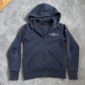 Mörkblå zipup hoodie från Peak Performance - Mörkblå hoodie från Peak Performance med broderad logga på bröstet. Tröjan har dragkedja, huva och två fickor framtill. Insidan är mjuk och fleecefodrad för extra komfort. Perfekt för kyliga dagar och en avslappnad stil. Däremot har armbågarna blivit lite uppslitna, där dragit ner priset enormt. Köpta för 1000, säljs för 200